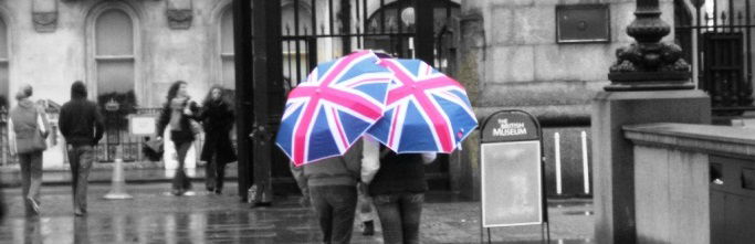 A couple of people stand in front of the British museum, hidden by huge Union Jack umbrellas.
