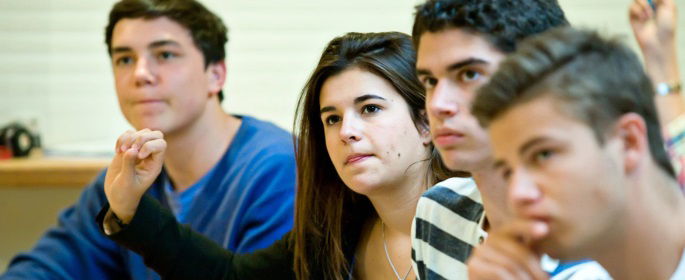 Image shows students raising their hands in class.