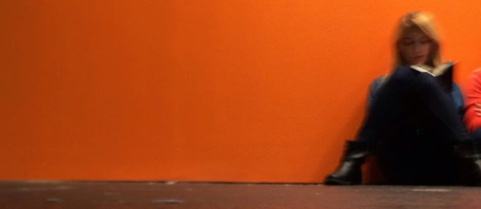 Image shows a woman reading a book, leading against an orange wall.