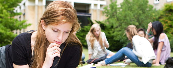 Girl reading book on grass with students in background
